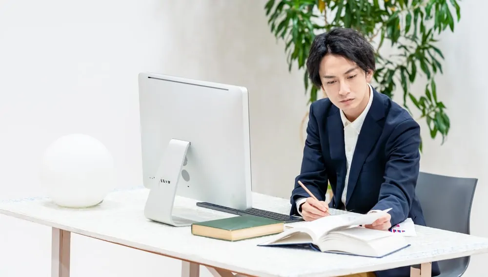 Man writing at desk with computer and books.